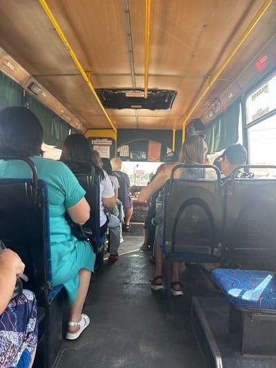 The image depicts the interior of a bus with several passengers seated. The bus has a wooden-paneled ceiling with yellow handrails. In the foreground, a woman in a turquoise dress is seated, her white sandals visible. To her right, a woman with long blonde hair is seated, wearing a grey tank top and shorts, and black sandals. Another passenger, a young person wearing a black baseball cap, is seated in front of her. Further down the aisle, other passengers are visible, some facing forward and others towards the rear of the bus. The seats appear to be upholstered in blue fabric with a red and blue pattern. Outside the windows, a sunny environment is visible, suggesting it is daytime. Text on the bus's interior includes "ПРОДАМ" (For Sale) and what appears to be a list of route numbers. The presence of "APTEЧКА" (First Aid Kit) suggests it might be a public transportation vehicle. The scene is consistent with the interior of a bus operating in the city of Obukhivka, Ukraine, given the Cyrillic text and general bus design common in the region.