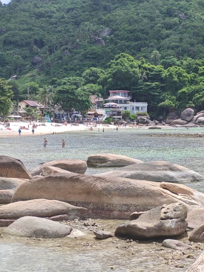 This media file captures a vibrant beach scene in Baan Chaweng Noi, Thailand. The foreground is dominated by large, rounded rocks partially submerged in clear, shallow water. The water reflects the bright daylight, creating a shimmering effect.
Beyond the rocks, a sandy beach is dotted with numerous people. Some are sunbathing on loungers, while others are walking or standing near the water's edge. A few individuals are wading in the shallow sea. The beach is lined with a collection of buildings, including what appears to be a multi-story white hotel or resort with red accents, and smaller structures with awnings, suggesting a lively tourist area. Trees, including palm trees, and lush green foliage form a dense backdrop, climbing up a steep, rocky hillside. The overall atmosphere appears relaxed and summery, indicative of a pleasant day. The time of day is likely midday, given the bright sunlight and shadows. The weather seems clear and sunny, ideal for a beach day. There is no visible text in the image.