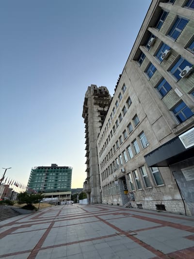 The image shows a wide-angle view of modern buildings and a pedestrian walkway in Shumen, Bulgaria. The perspective is from a low angle, looking up at the structures, and the image is rotated 90 degrees clockwise.

The primary subjects are two multi-story buildings. The building in the foreground is a light-colored, concrete structure with many windows and visible air conditioning units. Its architecture appears to be from the mid to late 20th century. To the right of this, a second building, partially covered in green netting, is under construction. This building is also multi-story and has a more contemporary design. The foreground also features a paved pedestrian area with a distinct pattern of reddish-brown lines on gray tiles. Several flags on poles are visible in the distance, near the construction site, suggesting a public or official area.

The scene is set in an urban environment, likely a plaza or a street with significant architectural elements. The sky is clear and a light blue, indicating fair weather. The lighting suggests it is either early morning or late afternoon, given the soft light and the absence of harsh shadows.

There are no visible people in the image. The overall mood is one of urban development and modern architecture. No significant text is legible in the image, though there are some signs on the buildings that are too small or indistinct to read.