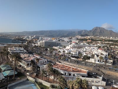 The image depicts an elevated, panoramic view of Los Cristianos, Spain, encompassing urban architecture, natural landscapes, and the coastline. Dominating the immediate foreground is a tennis court, partially visible in the lower left corner. Palm trees are scattered throughout the scene, particularly along the roadways and amongst the buildings. The buildings are primarily white with some orange and brown roofs. A busy road with car traffic runs horizontally across the middle ground. Beyond the urban sprawl, mountains loom in the background, partially obscured by clouds. The sea and a stretch of sandy beach are visible on the left side of the image. The sky is clear and blue.