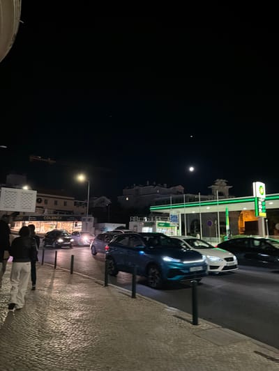 The nighttime image shows a street scene in Cascais, Portugal. A busy street with several cars driving on it. The cars are illuminated by streetlights and the headlights of other vehicles. On the right side of the image is a BP gas station with bright green and yellow signage. A sidewalk with cobblestone paving runs along the left side of the street, with several people walking on it. In the background, buildings are visible, some with lights on. The sky is dark with a moon and a few stars visible. The overall impression is one of an active urban environment at night.