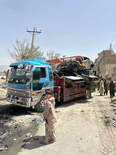 A blue Hino truck is parked on a street in Quetta City Tehsil, Pakistan. On top of the blue truck is a burnt vehicle being lifted by a red UNIC crane. A young boy in a tan outfit stands next to the truck. Several people are in the background standing in a group by a building. The street has debris scattered across it. There is a utility pole in the foreground.