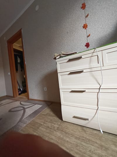 The image depicts an interior scene, likely a residential bedroom or living area, in the city of Tyumen, Russia. In the foreground, a light-colored dresser with multiple drawers is visible. A white cord, possibly a charger or cable, hangs down from the top of the dresser. To the left of the dresser, a portion of a patterned rug is visible on a wooden floor. Further to the left, a doorway with a wooden frame leads into another room, where some clothing and a vacuum cleaner can be seen. The walls are covered with textured wallpaper. A string of decorative orange leaf shapes hangs from the upper portion of the wall. There are no people clearly visible in the image.