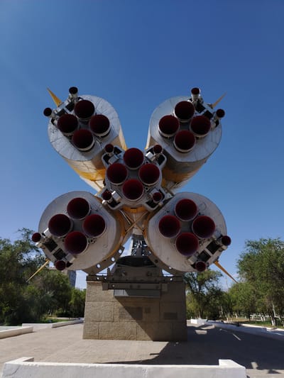 The image shows the base of a rocket, likely a monument, displayed outdoors in a public space. The rocket appears to be an older model, with multiple engine nozzles visible. It is mounted on a concrete pedestal, surrounded by a paved walkway and some greenery. The sky is clear and blue, suggesting a sunny day. The overall setting suggests a park or memorial site in Baikonur, Kazakhstan, a city known for its spaceport. No people are visible in the image.