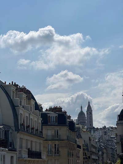 The image shows a view of Paris, France, including the Sacré-Cœur Basilica on Montmartre. The basilica's dome and tower are visible among the rooftops of the surrounding buildings. The foreground consists of Parisian architecture, with buildings featuring multiple stories, windows, and chimneys. The sky is partly cloudy with blue sky peeking through.