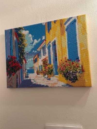 This media file is a close-up, angled shot of a diamond painting depicting a scene of a building facade with windows and flowering plants. The painting is made of small, colored beads that create a mosaic-like image. The dominant colors are yellow, blue, white, and various shades of green, red, and purple for the foliage and flowers.
The scene appears to be outdoors, possibly on a sunny day given the bright yellow and blue tones which suggest sunlight and a clear sky. The architecture is rendered with white and yellow horizontal and vertical lines, creating the impression of windows, balconies, or architectural elements. Lush green plants with clusters of red and purple flowers are visible, some appearing to hang from planters or windowsills. The bottom of the painting shows a darker blue area that could represent water or a shaded ground level, with more green foliage and red flowers. The painting itself is mounted on a wall with a textured, light-colored surface, and is slightly tilted, casting a subtle shadow.
There is no discernible text in the image. The overall impression is a vibrant, somewhat abstract representation of a picturesque building with abundant flora. The location context of Oława, Poland, doesn't provide specific visual cues within the painting itself, but suggests a European setting.