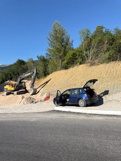 An outdoor daylight scene shows a construction site in Prijevor, Montenegro, under a clear blue sky. In the foreground, a dark grey asphalt road transitions to a light-colored gravel area. To the right of the gravel, a dark blue hatchback car is parked with its driver's side door and rear hatch open. The car is positioned next to a curb that separates the gravel from the asphalt.

Behind the car, a steep, beige-colored dirt embankment rises, dotted with sparse vegetation and some red pipes protruding from the ground. Further to the left, a large yellow excavator is parked on a mound of light brown sand and gravel. A dark grey Volvo excavator is positioned behind the yellow one, with its arm extended. Beyond the construction site, a dense forest covers the hillside, with mountains visible in the distance. The overall impression is that of an active construction project in a semi-rural or mountainous area. There are no people visible in the image.
