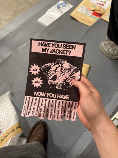 A person's hand is prominently featured, holding up a distinctive flyer designed with a black background and pink text/graphics. The flyer's left side asks, "HAVE YOU SEEN MY JACKET?", while the right side states, "NOW YOU HAVE". A detailed illustration of a jacket occupies the center. Below the jacket are two star-shaped logos; one contains the Greek letter 'ρη' and the other a stylized emblem. Along the right edge of the flyer are numerous tear-off tabs, each printed with the repeated phrase "NOT JUST UNIFORM."
The scene appears to be an indoor setting, likely a casual or creative space in London, UK. The floor is a neutral grey, possibly concrete. In the background, various items are visible: a brown cardboard parcel with labels, a pair of light blue sunglasses resting on white paper, and glimpses of two other individuals. One person is seen wearing dark pants and light-colored sneakers, while another wears grey jeans and a brown shoe. A yellow strip in the bottom right corner displays the partial text "The Graphical." The overall atmosphere is informal, suggesting the flyer is being shown or distributed.