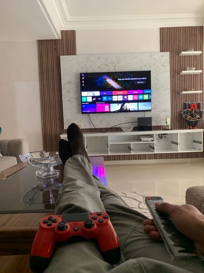 A person sits with a red PlayStation controller and a remote control in their hands, facing a TV screen displaying a user interface. Their legs are crossed, with socks on the feet resting near a glass dish on a table. The TV is mounted on a marble panel. Behind the TV, there is a bookshelf with empty shelves and decorations on the wall. The room appears to be a living space with light-colored walls and furniture.
