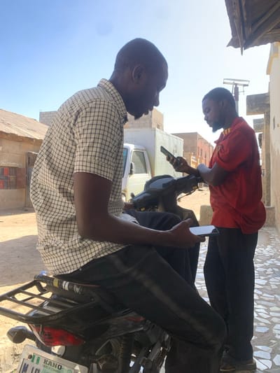 Two adult males are depicted outdoors in Gwagwarwa, Nigeria, under clear daylight. The male on the left, wearing a white and black checkered short-sleeve shirt and dark pants, is seated on a dark motorcycle. He holds a white smartphone horizontally in his left hand, looking downwards at its screen. The motorcycle has a black seat, a rear rack, and a partially visible Nigerian license plate from "KANO CENTRE OF COMMERCE." The male on the right, wearing a red short-sleeve shirt and dark pants, stands alongside the motorcycle. He holds a dark smartphone vertically with both hands, focusing on its screen. In the background, a white boxy vehicle is parked near several buildings, including one constructed from concrete blocks and another with a corrugated metal roof. The ground surface in the immediate foreground right is composed of irregular light-colored paving.