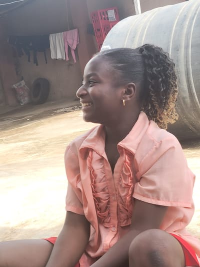 A young woman is outside, likely in a residential area of Madalla, Nigeria, based on the presence of a water storage tank, discarded tire, and laundry line. The woman, possibly a teenager, has a shaved head with curly hair extensions. She's wearing an peach colored frilly button up shirt with red shorts. She is crouching. The environment includes a water storage tank, laundry hanging on a clothesline, a tire, and general debris. The foreground shows sunlit pavement or ground.