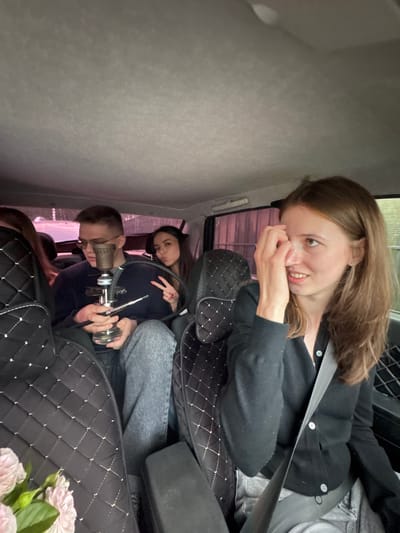 The media file shows three people in what appears to be the back seat of a vehicle. In the foreground, a woman with long brown hair is looking at the camera with a playful expression, her hand near her face. She is wearing a black cardigan over a black top. Behind her, another woman with dark hair is visible, making a peace sign with her fingers. She is wearing a dark blue top. In the center, a person with short hair and glasses, wearing a dark top, is holding a hookah. The interior of the car is visible, with quilted black seat covers. In the bottom right corner, there is a bouquet of light pink flowers. The overall mood seems casual and relaxed. The lighting suggests it could be daytime.