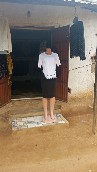 The image shows a storefront in Kuru Dungus, Nigeria. A mannequin stands in front of an open doorway, displaying a white short-sleeved shirt with a decorative hem and a black skirt. The doorway leads into a shop filled with clothing. To the right of the doorway, a black shirt with a white pattern hangs from a hook outside the building. The building's exterior is made of rough, light-colored plaster. The ground in front of the store is dirt. The scene appears to be outdoors, with natural lighting.