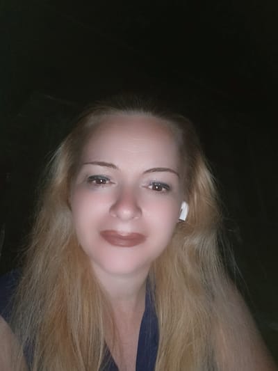 This is a close-up, selfie-style photograph of a blonde woman with fair skin and light brown lipstick. She is wearing a dark blue garment and has a white wireless earbud in her right ear. The background is dark and appears to be indoors, with some textured surface visible. There is no indication of the location within the city of Chernihiv, Ukraine.