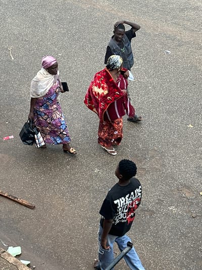 In Bukuru, Nigeria, four people are visible on a textured, asphalt surface. In the foreground, a young male with dark, curly hair wears a black t-shirt with red and white graphic text and light blue jeans. He is walking away from the camera, holding a dark metal pole. Behind him and slightly to the left, a woman with a pink headscarf and a floral patterned dress, carrying two bags, walks to the left. To her right, a woman in a red patterned garment is walking forward, and a man in a dark vest over a t-shirt and dark pants is standing with his right hand on his head. The environment is a street with scattered debris including a piece of wood and some plastic items.