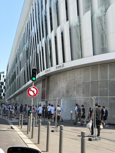 This image captures a scene outside a modern building with a distinctive facade of vertical silver fins and curved glass panels, located in Nanterre, France. A crowd of people is gathered, some standing in a queue behind a temporary metal barrier. The barrier runs parallel to the building's entrance, which is marked with signs indicating "Entrée 23" and "Entrée 21". A traffic light shows a green signal, and a circular traffic sign with a red border and a diagonal line through a left-pointing arrow is mounted on a pole next to it. The pavement in the foreground has bollards. Some individuals are wearing white t-shirts, and others are carrying bags. The sky is clear and blue.