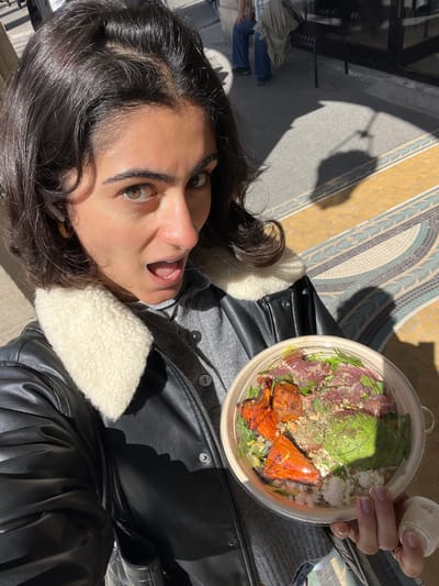 A woman takes a selfie while holding a bowl of food, possibly salad, in the city of Paris, France. The woman is wearing a grey shirt and a black leather jacket with a white wool lining. She has dark curly hair, fair skin, and wide eyes. Her mouth is open in a surprised expression. The bowl of food appears to contain lettuce, avocado, some red meat or vegetables, and cheese. The background shows a mosaic floor, with shadows falling across it. There is also a wrought iron railing visible in the background.