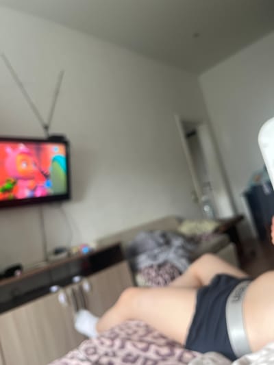 The image depicts an indoor scene, possibly a bedroom, with a television on the wall displaying a cartoon. A person is lying down on a bed in the foreground, wearing black shorts and white socks. The environment appears to be a typical living space with furniture such as a cabinet and a couch visible in the background. There is no identifiable information within the image that would allow for its location to be specified as Chelyabinsk, Russia.