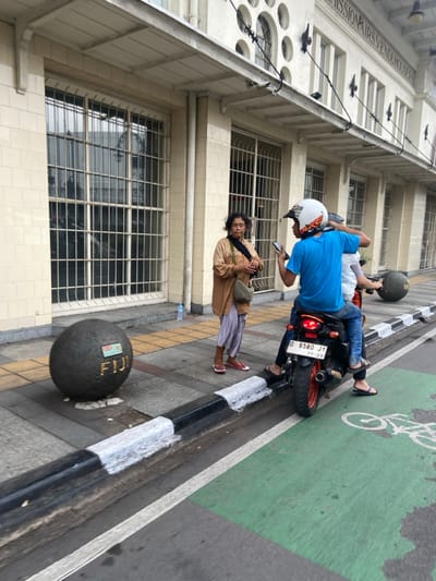 The scene is a city street in Bandung, Indonesia, located at the coordinates -6.922 latitude and 107.61 longitude. A woman wearing a brown jacket, long, dark skirt, and sandals stands on the sidewalk. She is facing a man wearing a white helmet and blue shirt riding a motorcycle. The motorcycle has a license plate that reads "D 6580 JY 11-29". The motorcycle is parked on a bicycle lane marked with a green painted surface. The man is holding a smartphone in his right hand and his left hand is on the motorcycle's handlebars. The woman is looking at the man. Behind the man on the motorcycle is another person, partially obscured from view, wearing a blue shirt. There is a large, gray, round object painted with the word "FIJI" on the sidewalk to the left of the woman. The building in the background is white, with large windows and a tiled facade. The sidewalk is gray, with a black and white curb separating it from the street. The street is asphalt. The scene is a public street. The time of day is daytime, as the sky is bright and the sun is shining. There is a small amount of shadow in the background.