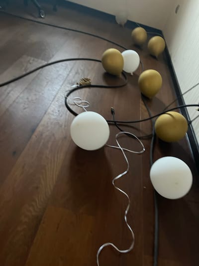 The image shows a string of globe lights on a wooden floor. The lights are a mix of gold and white, and they are connected by a black cord. Some of the lights appear to be deflated or damaged. The scene is lit by natural light, and there are shadows cast on the floor. There are no people or animals visible in the image. The location is the city of Orekhovka, Russia.