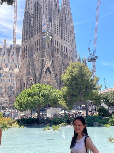 The image shows a woman standing in front of the Sagrada Familia in Barcelona, Spain. The Sagrada Familia is a large, unfinished basilica designed by Antoni Gaudí. The woman is smiling and wearing a white tank top. Behind her, the Sagrada Familia towers over a park with trees and a light blue pool of water. Cranes are visible behind the basilica, indicating that construction is ongoing. A crowd of people can be seen in the background, some sitting and some standing. The sky is clear and blue.