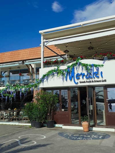 This image depicts the exterior of "Meraki Greek Food & Orient Grill," a two-story restaurant located in Varna, Bulgaria, as indicated by "Meraki Varna" stenciled on the pavement. The building's ground floor features a white facade with brown-framed glass doors and windows, displaying the restaurant name in large blue script with "Greek Food & Orient Grill" in smaller black text. Artificial green vines with purple wisteria-like flowers are draped decoratively above the entrance. Potted green plants, resembling oleander, are placed on the concrete pavement outside. The upper level includes an open-air terrace with tables and chairs, a glass balustrade, and red flowers in planters. A white retractable awning system extends over this area, with string lights suspended beneath. Behind the upper terrace, a building with a terracotta-tiled roof is visible under a clear blue sky with sparse white clouds. The concrete pavement in the foreground features partial white painted markings and a blue oval graphic containing "Meraki Varna" along with what appears to be "Love" and a Greek flag icon. Reflections in the glass doors and windows show glinting water, possibly a harbor or sea, and faint outlines of human figures and a ship's mast. No individuals are directly visible.