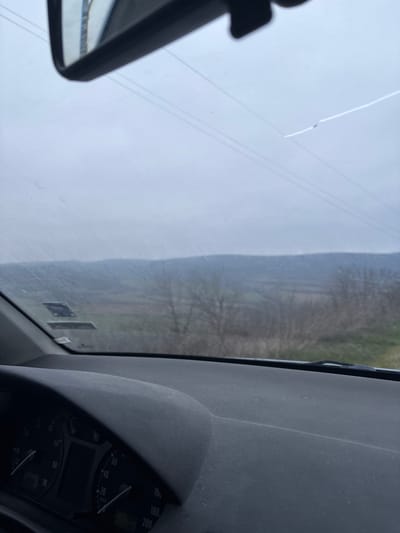 The scene depicts a view from the interior of a vehicle, looking outwards through a dirty windshield. The presumed location is Balkantsi, Bulgaria.
The upper portion of the image shows an overcast, light grey sky. Several dark, horizontal lines, consistent with power or telephone lines, traverse the sky. A prominent crack is visible in the upper right quadrant of the windshield, originating from a small impact point and extending horizontally. A rearview mirror is partially visible at the top center, reflecting a light source or interior structure.
Below the sky, a distant horizon line of dark, possibly forested hills or mountains is visible, partially obscured by atmospheric haze. In the midground, various fields, some green and some brown, are discernible. Bare-branched trees and shrubs are scattered across this midground. To the far right, a patch of green ground suggests a roadside verge or field edge.
The lower portion of the image reveals the vehicle's interior. The dark grey dashboard, appearing dusty, extends across the lower frame. The top of the instrument cluster is visible on the lower left, displaying dark circular dials with white markings, including a speedometer and tachometer, both indicating low or zero readings. No individuals are visible within the frame.
