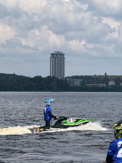 An aerial shot captures a jet ski race on the Kama River in Perm, Russia, on a cloudy day. The foreground features a jet skier in a blue and white helmet and a blue racing suit, identified by the number "1" on their back, riding a green and black jet ski with the number "91" displayed prominently. The jet ski is creating a spray of water as it moves. In the mid-ground, a dense line of dark green trees borders the river, beyond which several buildings are visible. A tall, modern skyscraper with a white facade and numerous windows stands out amongst these structures. The background is dominated by a cloudy sky. Another jet skier, wearing a yellow and black helmet and a blue racing suit with the number "2" visible, is partially visible in the lower right corner of the frame, looking towards the race. The overall scene suggests an outdoor sporting event taking place on a body of water with an urban backdrop.