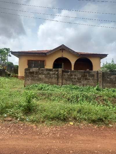 The image shows a single-story, yellow house with a brown roof. The house is located in Ita Oniyan, Nigeria, at coordinates 7.291 latitude and 5.166 longitude. The house has two arched doorways and a single window. The house is surrounded by a low concrete wall. In front of the wall, there is a patch of green grass, and in the foreground, there is a dirt road. The sky is overcast with white clouds. There are no people visible in the image. The scene is taken from a perspective slightly elevated from the ground, giving the impression that the viewer is standing on the dirt road in front of the house. There are multiple electric power lines running across the sky. The image was likely taken in the afternoon, as there are no shadows visible.
