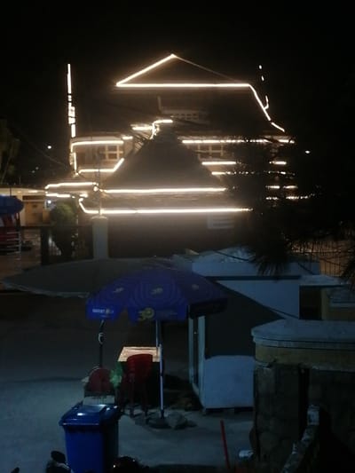 In Da Lat, Vietnam, a nighttime scene unfolds with a building adorned with bright white decorative lights outlining its roofline and structure. In the foreground, a blue trash bin sits on the pavement, accompanied by a red chair and a small table under a blue umbrella. Behind the table, a white kiosk-like structure is visible. The environment appears to be an urban street, with the building's illumination casting a glow against the dark night sky.