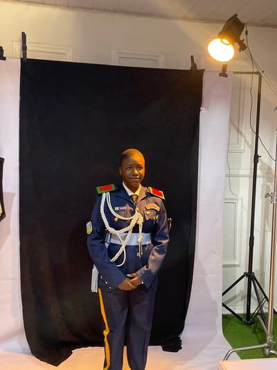 A photograph features a young person in a navy blue military-style uniform with gold accents, likely taken in a studio or indoor setting in the city of Barakin Akawo, Nigeria. The subject stands against a backdrop comprised of white and black fabric. Lighting equipment is visible in the background, including a light fixture on a stand. The subject's uniform displays decorative white ropes and insignia. The environment seems like a controlled photography space.