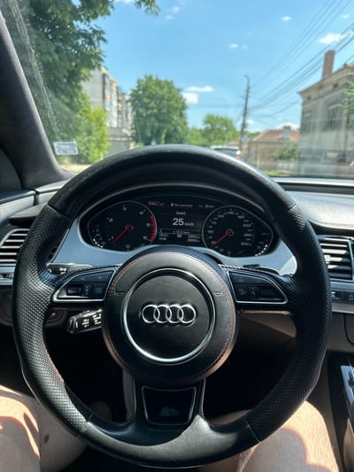 This media file shows a close-up view of the interior of an Audi car, focusing on the steering wheel and dashboard. The steering wheel is black leather with perforated grips and red stitching. The Audi logo is prominently displayed in the center of the wheel. On the dashboard, a digital display shows the speed as 25 km/h. A traditional speedometer with a red needle is also visible, pointing to the 25 km/h mark. Other indicators on the dashboard include temperature (30.5℃) and time (14:46).

Through the windshield, the scene outside appears to be a residential area on a sunny day. Buildings with brick accents and trees are visible. The sky is bright blue with scattered clouds. The presence of overhead electrical wires suggests an urban or suburban setting. The lighting indicates it is daytime, likely afternoon given the time displayed on the dashboard. A person's leg and knee are visible in the lower right portion of the frame, suggesting the driver is operating the vehicle. The overall impression is a snapshot of everyday driving.