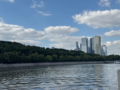 This is a wide-angle, horizontal, eye-level shot of a wide river in Moscow, Russia, on a partly cloudy day. The water in the foreground is choppy and reflects the sky and clouds. In the middle ground, a concrete embankment separates the river from a densely wooded hillside covered in lush green trees. Beyond the trees, a modern cityscape of tall skyscrapers rises against the blue sky. The buildings are a mix of glass and metal, with some featuring distinctive architectural designs. Two boats are visible on the river in the distance, one appearing to be a ferry. The sky is a vibrant blue with scattered white, fluffy clouds. The overall mood is serene and picturesque, highlighting the contrast between the natural landscape and the urban environment. The time of day appears to be midday or early afternoon. There is no visible text in the image.