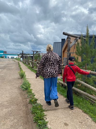 In Votkinsk, Russia, under a cloudy sky, two individuals walk away from the camera on a concrete path. The person in front, wearing a leopard-print jacket and bright blue pants, walks on the left side of the path. To their right, a younger person, in a red jacket and dark pants, walks slightly ahead, touching a wooden fence. The path is bordered by grass and wildflowers. Behind the fence, a modern building with wooden accents and a dark roof is visible, along with a pine tree. In the background, a street with buildings and power lines suggests an urban or suburban setting.