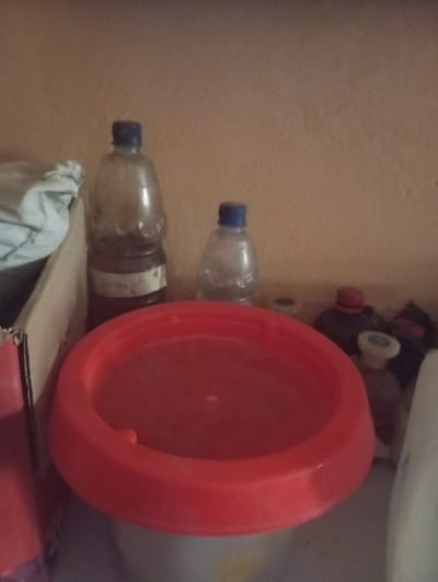 The image shows a collection of items on a shelf against a textured, peach-colored wall. In the foreground, a large, bright red, circular basin or lid is prominently displayed. Behind it, to the left, is a tall, clear plastic bottle with a blue cap, containing a dark liquid and a label. To the right of this bottle, a smaller, clear plastic bottle with a blue cap is visible. Further to the right, there are several other objects including a bottle with a red cap, and a white object that could be a bottle or container. A cardboard box is partially visible on the far left. The scene appears to be an indoor storage area, possibly a shelf in a home or shop, located in the city of Jalingo, Nigeria.