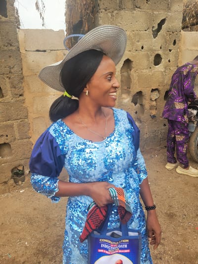 A woman in a light blue sequined dress and a wide-brimmed straw hat is standing outside in Madalla, Nigeria. She is holding a blue tote bag labeled "CONGRATULATIONS ON YOUR INDUCTION/OATH TAKING FROM SCHOOL OF HEALTH TECHNOLOGY GULEJA (SOHITE)." She is smiling and looking to her left. In the background, a person wearing a purple patterned outfit and white shoes is visible. The surrounding environment includes a brick structure and dirt ground.