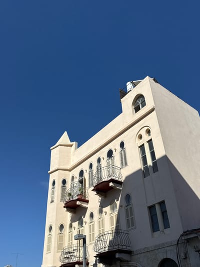 An upward-angle shot shows the cream-colored facade of a building against a vibrant blue sky in Tel Aviv, Israel. The building features arched windows, some with shutters, and ornate balconies with black metal railings. A small tower with a pointed roof rises from the left side of the building, and a water tank is visible on the roof. The building's architecture suggests a blend of styles, possibly influenced by Ottoman or Art Deco design, common in Tel Aviv's older neighborhoods. A shadow from the building stretches across its facade, indicating a bright sunny day. The lower part of the building shows stone masonry. The scene is devoid of people.