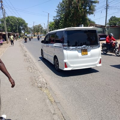 A street scene in Nairobi, Kenya, is depicted under a clear blue sky. A white Toyota Noah minivan, license plate KDW 903Q, is positioned on an asphalt road, facing away from the viewer. The vehicle has tinted rear windows, a chrome strip above the license plate, and a rear spoiler. To the right of the minivan, a person wearing a red jacket and a black helmet rides a red motorcycle, traveling in the same direction. Multiple other motorcycles with riders are visible further down the road, some with yellow-jacketed operators. On the left, an unpaved dirt and grass shoulder borders the road, alongside which rudimentary roadside stalls with corrugated iron roofs are visible. A male pedestrian in a light green shirt and dark trousers walks on this shoulder, moving away from the camera. Overhead utility lines crisscross the frame, supported by wooden poles along the left side of the road. The lower left foreground shows a partial view of a dark-skinned arm and hand with a white shoe. Trees and various structures are present in the background, including a building on the far right with what appears to be discarded furniture.