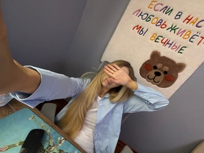 This is a vertical, eye-level, full shot photograph of a young woman lying down and covering her face with her hands. She has long, blonde hair that is spread out across the frame. She is wearing a light blue button-down shirt over a white tank top and light blue pants. The woman is lying on a light-colored rug with a cartoon bear motif. Below the bear, there is text embroidered in various colors. The visible text reads "ЕСЛИ В НАС ЛЮБОВЬ ЖИВЁТ, МЫ ВЕЧНЫЕ". This translates from Russian to "If love lives in us, we are eternal." The scene appears to be indoors, possibly in a bedroom or living area, with a desk visible in the background. The overall mood is relaxed and contemplative, with the woman's gesture suggesting a sense of privacy or perhaps shyness. The lighting is soft, indicating it could be daytime, although there are no clear indications of time or weather. The presence of a mousepad with a floral design on the desk, and the general domestic setting, hint at a casual, personal space in Moscow, Russia.