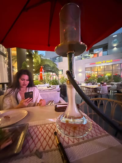 A woman with long dark hair is sitting at a table in an outdoor cafe, looking at her phone. She is wearing a white lace top. In front of her is a hookah, with smoke rising from the glass base. The table is covered with a light-colored tablecloth with a red diamond pattern. On the table are plates, cutlery, and a glass.
The background shows a street scene at night. Buildings with illuminated windows and signs are visible. Some of the signs appear to be in Vietnamese, with text like "COM & PHO" and "VIETNAMESE CUISINE". There are also palm trees and outdoor seating areas with wicker chairs and couches. Lights are strung across the scene, suggesting a festive atmosphere. The overall setting appears to be a commercial area, possibly a restaurant or entertainment district. The time of day is evening or night, indicated by the artificial lighting. The weather appears to be pleasant, as people are dining outdoors. The woman's expression is neutral as she focuses on her phone.