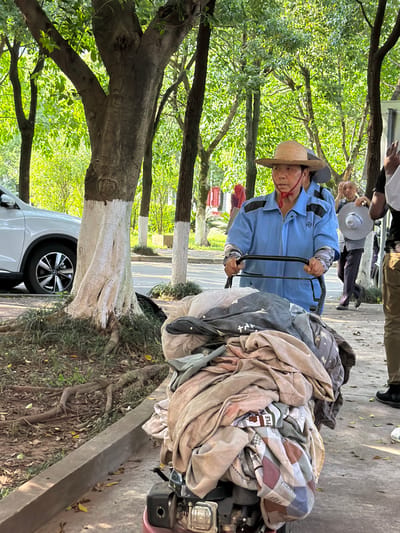 A man in a blue uniform and straw hat is pushing a cart filled with blankets down a sidewalk in Jiangjin, China. The area has trees and a parked car nearby. Other people are visible in the background.