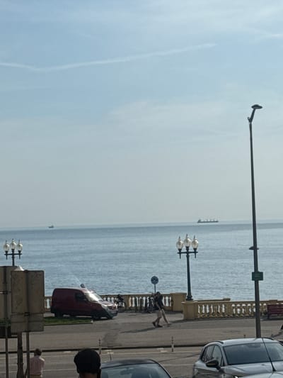 The image depicts a street scene in Porto, Portugal, with the Atlantic Ocean in the background. A body of water stretches to the horizon, where a large cargo ship is visible. The foreground shows a street with vehicles, including a red van, and pedestrians walking along a promenade. Decorative lampposts line the promenade, and a balcony with ornate railings separates the street from the sea. The sky is clear with a few wispy clouds. The overall impression is a bright, sunny day in a coastal city.