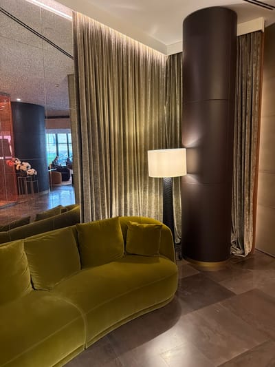 This image captures an interior scene featuring a plush, olive-green sofa in the foreground. Behind the sofa, a tall, dark brown cylindrical column stands prominently, draped with textured, golden-brown curtains. To the left of the column, a floor lamp with a white lampshade casts a warm glow. The background includes a large mirrored wall reflecting a seating area where two individuals are visible, one seated and looking at a laptop, the other in the distance. The floor is tiled with large, dark brown slabs. The environment appears to be a hotel lobby or a similar upscale public space, possibly in Saint Petersburg, Russia, given the context provided.