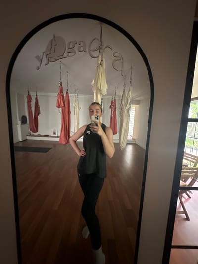 A young woman is standing in a room, taking a selfie in a mirror. The room appears to be a studio or fitness space, with several aerial yoga hammocks hanging from the ceiling. The floor is made of wooden planks. "Dream" is written on the mirror. The image does not provide information regarding Esenyurt, Turkey.