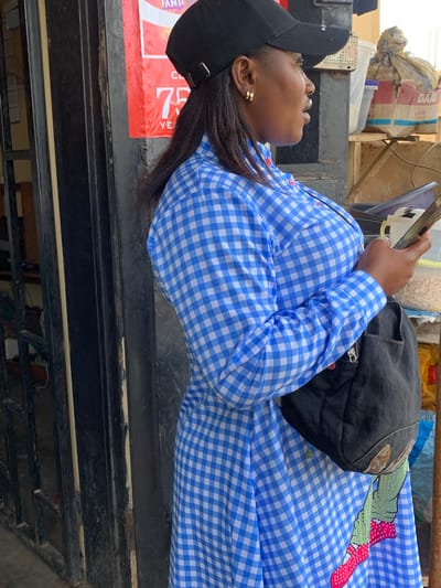 In Barakin Akawo, Nigeria, a woman wearing a blue and white checkered dress, a black baseball cap, and gold hoop earrings stands outdoors, partially obscured by a metal gate. She is holding a smartphone in her right hand and has a dark-colored bag slung over her shoulder. Behind her, a red poster with white lettering is visible, along with shelves stocked with various items, including a bag of Dano milk powder and plastic containers filled with what appear to be grains or nuts. The environment suggests a commercial setting, possibly a shop or market stall.