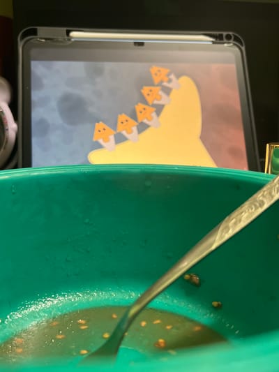 The image depicts a close-up view of a green bowl containing some food and a spoon. In the background, a tablet displays an animation of several orange triangular characters with faces. The scene appears to be indoors, possibly in a domestic setting. There is no identifiable information in the image that would suggest a specific location in Sevastopol, Ukraine.
