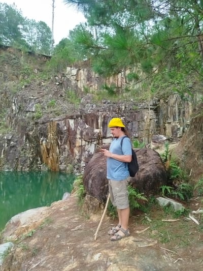 A person wearing a yellow hat, blue t-shirt, and khaki shorts stands on the edge of a quarry in Lán Tranh, Vietnam. The person is looking at a phone and holding a stick. A large rock is behind them, and a backpack is on their back. The quarry has steep, rocky walls and a body of green water at the bottom. Pine trees are visible in the background.