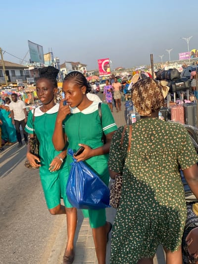 Two women in green uniforms stand on a street with a blue plastic bag. One woman holds a phone. Many people and luggage are nearby, including a woman in a green dress. Buildings and billboards are visible in the background under a clear sky. The location appears crowded and bustling.