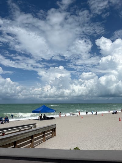 The image shows a beach scene in Englewood, United States. A wooden walkway leads down to the sandy beach. Several people are visible on the beach, some are sitting under a blue canopy, while others are near the water. The ocean has moderately sized waves and the water is a greenish hue. The sky is partly cloudy with large, white cumulus clouds dominating the upper portion of the frame. A rescue board is propped up on the sand near the water's edge. Orange traffic cones are placed sporadically on the beach.
