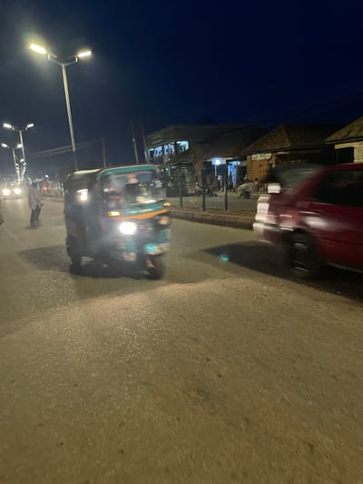 In Bukuru, Nigeria, at night, a teal and yellow auto-rickshaw is captured in motion on a paved road under streetlights. To the right, a blurry red vehicle is also moving, its headlights casting light on the road. In the background, several buildings with illuminated signs line the street, suggesting a commercial area. A person in a light-colored garment is standing on the left side of the road, facing the moving vehicles. The overall scene depicts a busy street at night, with vehicles and pedestrians present.